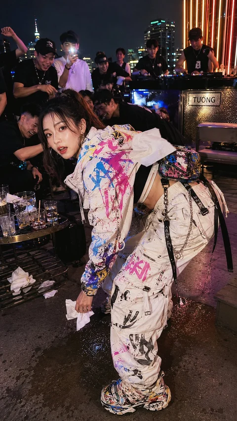 A person in colorful printed streetwear leans forward beside a busy outdoor bar at night.