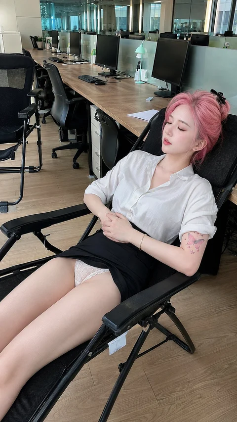 A person with pink hair reclines in an office chair beside a long desk of computer monitors.